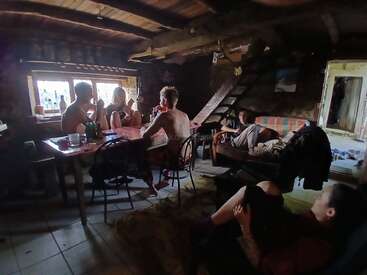 Five people relax in a rustic, dimly lit cabin. Three sit at a table near a window, two lounge on couches, enjoying a cozy, casual atmosphere.