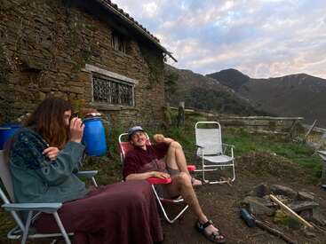 Two people relax in chairs outside a rustic stone house, surrounded by mountains. One drinks from a cup, while the other smiles, enjoying the peaceful nature.