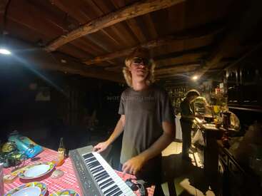 A man with long hair and sunglasses plays a keyboard in a dimly lit rustic kitchen, while another person prepares food in the background. Cozy atmosphere.