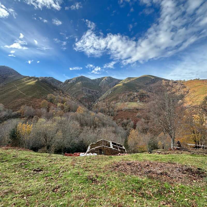 A rustic stone building sits in a grassy field, surrounded by autumn trees, under a dramatic, partly cloudy blue sky, with rolling, green mountains behind.