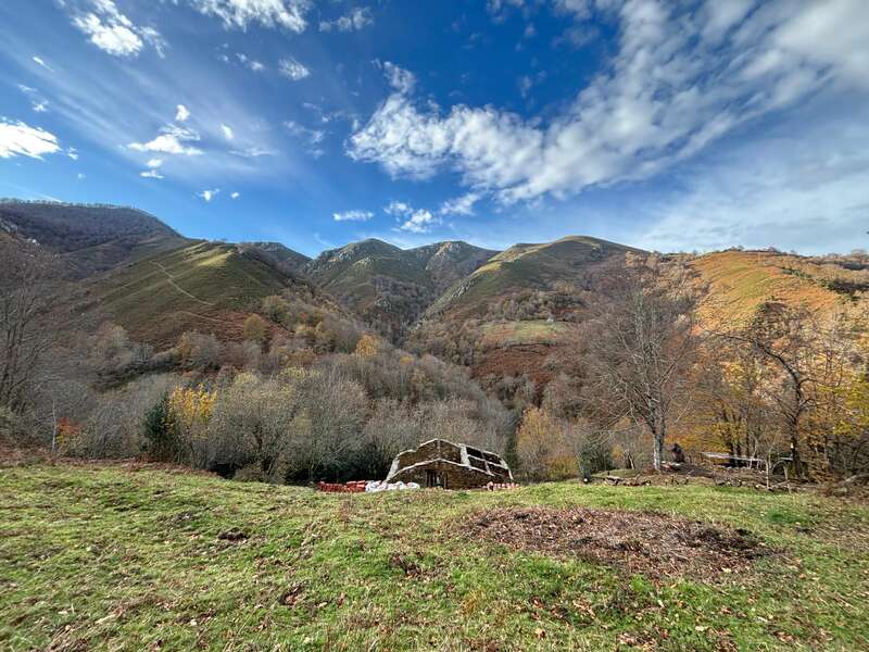 A rustic stone building sits in a grassy field, surrounded by autumn trees, under a dramatic, partly cloudy blue sky, with rolling, green mountains behind.