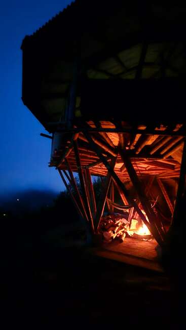 Eine Bambuskonstruktion, die in der Abenddämmerung von einem glühenden Lagerfeuer beleuchtet wird. Der tiefblaue Himmel unterstreicht den Kontrast zwischen dem warmen Feuerlicht und der Nacht. Gemütliche Atmosphäre.