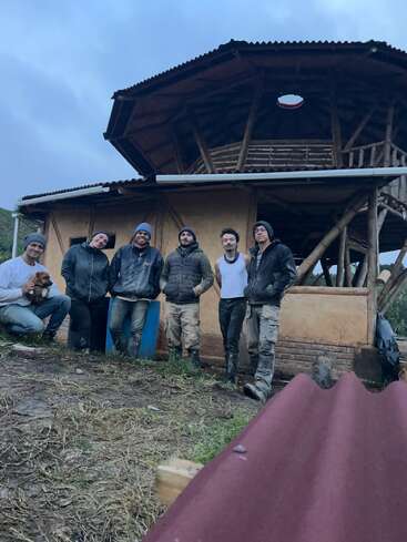 Sechs Personen posieren vor einem rustikalen, zweistöckigen, runden Holzhaus. Sie tragen Arbeitskleidung, wirken glücklich und eine Person hält einen kleinen Hund.