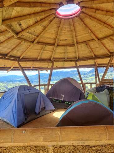 Mehrere Zelte sind unter einem runden Bambusdach mit einem Dachfenster aufgebaut. Draußen sind wunderschöne Berge und der blaue Himmel zu sehen, was eine gemütliche, rustikale Camping-Atmosphäre schafft.