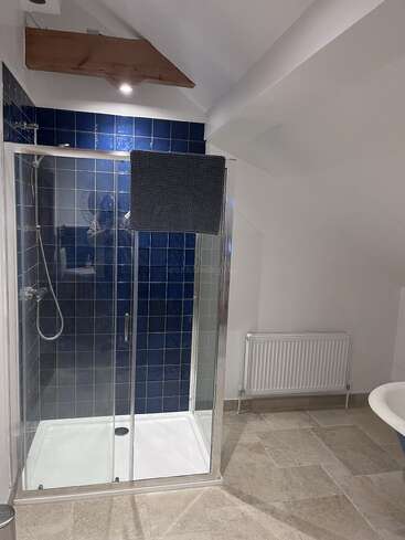 Dieses Badezimmer hat eine Glasdusche mit blauen Fliesen, einen Heizkörper, einen beigen Fliesenboden, weiße Wände, einen dunklen Handtuchhalter und eine coole schräge Holzdecke.