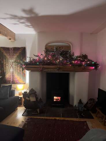 Ein gemütliches Wohnzimmer mit einem brennenden Kamin, einem geschmückten Kaminsims mit Girlanden und bunten Lichtern, einem Spiegel, sanfter Beleuchtung, Wandteppichen und einer einladenden Sitzecke.