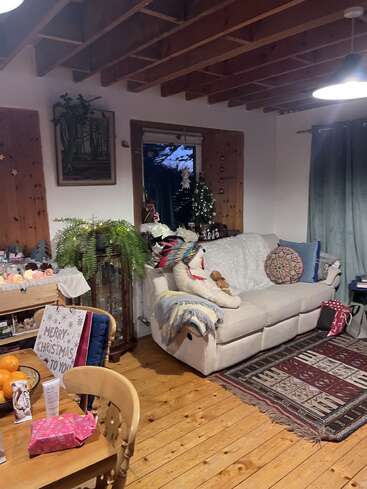 Ein gemütliches Wohnzimmer, das für Weihnachten geschmückt ist, mit einem kleinen Baum, Geschenken, einem bequemen Sofa, Zimmerpflanzen, Holzböden, freiliegenden Balken und warmer, einladender Beleuchtung.