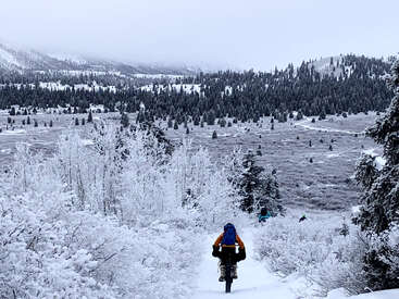 Eine Person fährt mit dem Fahrrad durch eine verschneite Winterlandschaft, umgeben von frostbedeckten Bäumen, sanften Hügeln und nebelverhangenen Bergen in der Ferne. Eine friedliche Szene.