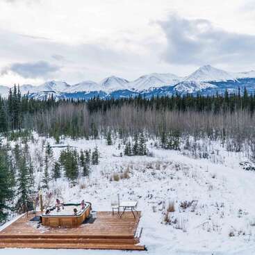 Vier Personen entspannen in einem Whirlpool auf einer Holzterrasse, umgeben von verschneiten Wäldern. Majestätische, schneebedeckte Berge und ein wolkenverhangener Himmel vervollständigen die ruhige Winterlandschaft.