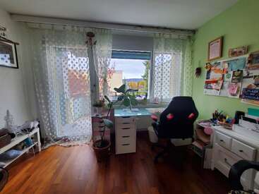 A cozy, well-lit study room with a large window, white desk, office chair, potted plants, bulletin board, hardwood floors, and green accent wall. Organized and inviting.