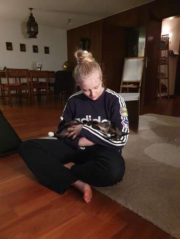 A blonde woman sits cross-legged on a wooden floor, gently cradling a cat in her arms. The setting is a cozy, warmly-lit living room.