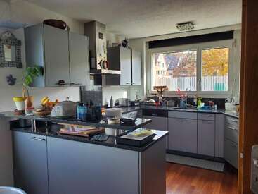 This kitchen features gray cabinets, black countertops, various appliances, and utensils. There's a window with an outdoor view, houseplants, fruits, and books on the counter.