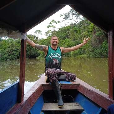 A smiling man sits cross-legged at the front of a boat, wearing a Boston Celtics jersey, surrounded by lush green jungle and calm river waters.