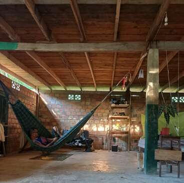 A man relaxes in a hammock inside a rustic brick-walled room with wooden ceiling, minimal furniture, plants, and sunlight streaming through small windows, creating warmth.