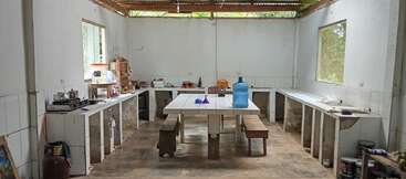 Essa é uma cozinha simples com armários abertos, uma mesa grande com bancos, um bebedouro, janelas, vários utensílios e itens básicos de cozinha à vista.