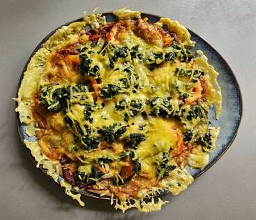 Una pizza casera en un plato azul, cubierta de queso fundido, espinacas, tomates y verduras varias, con los bordes de queso crujiente desparramándose por la corteza.