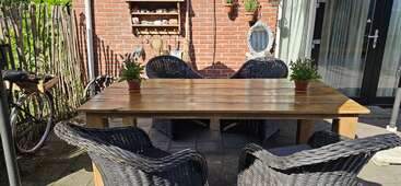 Una mesa de madera para exteriores con cuatro sillas de mimbre y dos macetas con plantas en un patio. La pared de ladrillo, la bicicleta, la decoración del jardín y la luz del sol crean un ambiente acogedor.