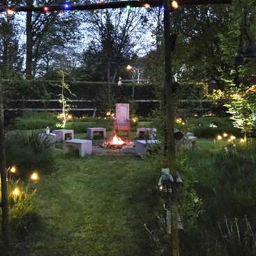 Un acogedor jardín al anochecer con una hoguera central, rodeada de bancos y una silla. Las luces de hadas y los farolillos crean un ambiente mágico y acogedor.
