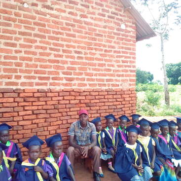The image depicts a group of children in graduation attire posing with an adult in front of a brick wall, likely celebrating a significant academic milestone.