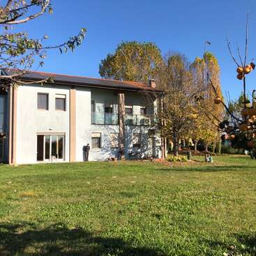 L'image représente une maison à deux étages avec un extérieur blanc, un toit marron et un balcon, située sur une pelouse avec des arbres et un ciel bleu clair.