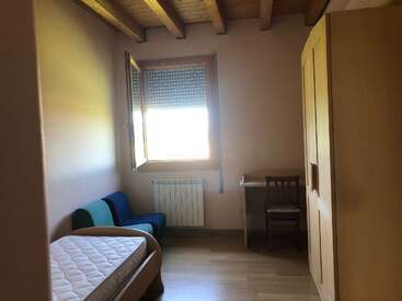 The image depicts a small, cozy room with a bed, a window, a desk, and a chair, featuring white walls and a wooden floor.