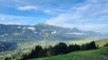 Um vale pitoresco com campos verdejantes, uma vila encantadora, florestas densas e montanhas imponentes sob um céu azul brilhante, pontilhado por nuvens e névoa.