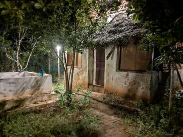 Uma casa antiga e rústica com telhado de palha e paredes desgastadas pelo tempo, cercada por plantas e árvores crescidas, iluminada por uma única luz externa à noite.