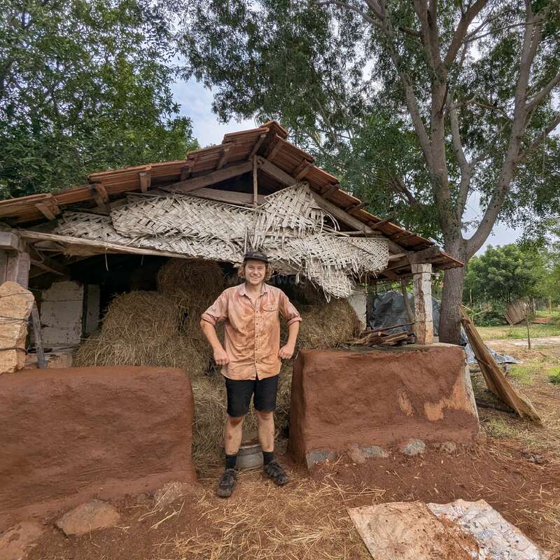 A person stands smiling in front of a rustic, thatched-roof barn with hay bales inside, surrounded by green trees and rural scenery, muddy and content.