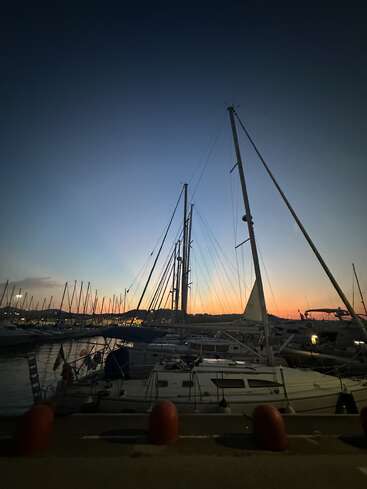 Sailboats are docked in a marina at dusk, silhouetted against a colorful sunset sky. Calm water reflects boats, creating a peaceful evening harbor scene.
