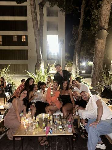 A group of friends gathers at night outdoors, smiling around a table filled with drinks, enjoying each other's company in a lively, festive atmosphere.