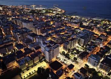 This image shows a coastal city at night, illuminated by streetlights. Buildings are densely packed, roads crisscross, and the shoreline stretches into the distance beside the sea.