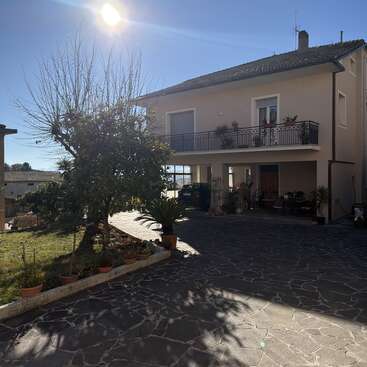 Ein zweistöckiges Haus mit einem Balkon, umgeben von Topfpflanzen und Bäumen, steht in der prallen Sonne und wirft Schatten auf die gepflasterte Auffahrt.