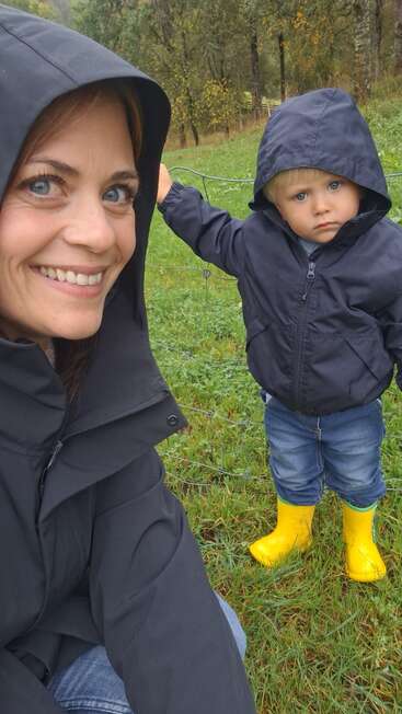Eine lächelnde Frau und ein ernst dreinblickendes Kind stehen im Freien, beide in schwarzen Regenjacken. Das Kind hat gelbe Stiefel an. Grünes Gras und Bäume umgeben sie.