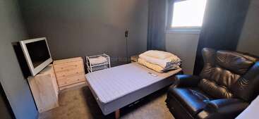 Dies ist ein kleines, gemütliches Schlafzimmer mit grauen Wänden, einem Einzelbett, einem schwarzen Ledersessel, einem Fernseher, Holzschubladen und einem Fenster mit Tageslicht.