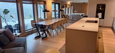 Esta imagem mostra uma moderna cozinha, sala de jantar e sala de estar em plano aberto com piso de madeira, janelas grandes, uma longa mesa de jantar, móveis contemporâneos e luz natural.