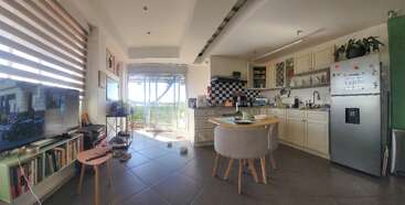 This image shows a cozy kitchen and living space, featuring a dining table, TV, bookshelves, large window with sunlight, plants, refrigerator, and colorful decor.