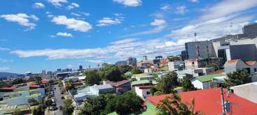 A vibrant cityscape under a bright blue sky with scattered clouds, showcasing colorful rooftops, leafy trees, modern buildings, and a lively urban environment. Busy, sunny day.