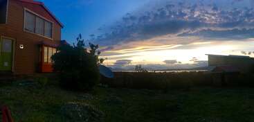 Um belo pôr do sol ilumina o céu atrás de uma casa e um jardim, com nuvens dramáticas, vegetação exuberante e atmosfera tranquila, criando uma cena pacífica e serena.