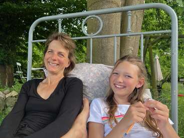 Eine Frau und ein Mädchen sitzen zusammen auf einer Bank im Freien, lächeln und lachen. Im Hintergrund sind Bäume und Grünflächen zu sehen, was eine friedliche und fröhliche Atmosphäre schafft.