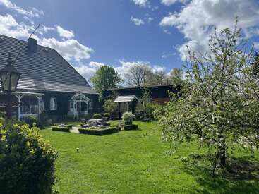 Ein charmantes Landhaus mit Ziegeldach, umgeben von üppigem Grün, blühenden Bäumen, Gartenmöbeln und strahlendem Sonnenschein unter einem blauen, bewölkten Himmel.