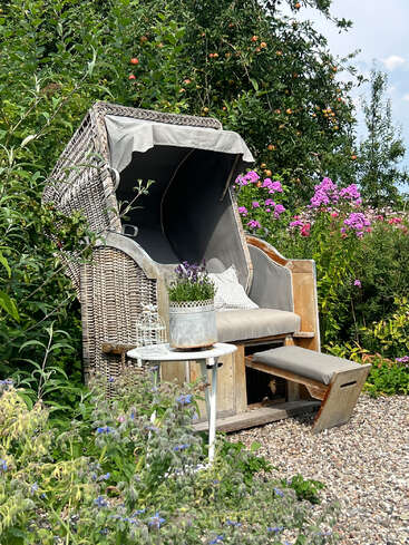 Ein gemütlicher Strandkorb aus Korbgeflecht mit Sonnendach steht in einem üppigen Garten, umgeben von blühenden Blumen, grünem Laub und einem kleinen weißen Tisch mit Deko.