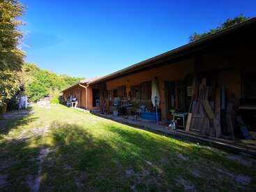 A imagem mostra um prédio comprido, de um andar, com telhado inclinado, situado em uma área gramada, cercado por árvores e um céu azul claro.