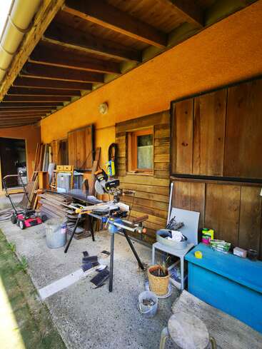 A imagem mostra uma área de oficina externa desordenada, com uma serra, um cortador de grama e várias ferramentas, situada do lado de fora de uma casa com revestimento de madeira e piso de concreto.