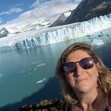 L'image montre une femme prenant un selfie devant un glacier et un lac, avec des montagnes en arrière-plan par une journée ensoleillée.