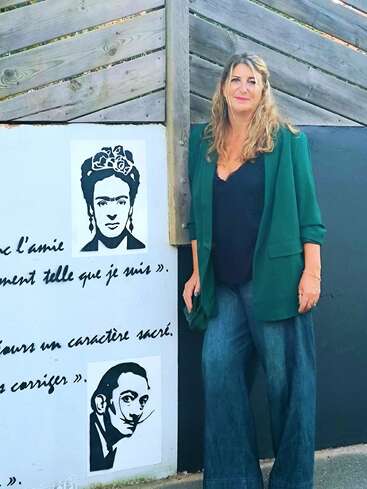 Uma mulher de cabelos ondulados usa blazer verde, blusa escura e jeans azul largo, ao lado de um mural com Frida Kahlo, Salvador Dalí e texto em francês.