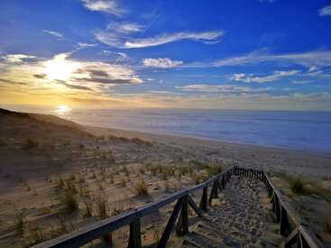 A luz dourada do sol banha a praia de areia. Escadas de madeira levam você até a costa tranquila, onde o céu azul encontra ondas suaves sob nuvens finas ao pôr do sol. Serenidade pacífica.