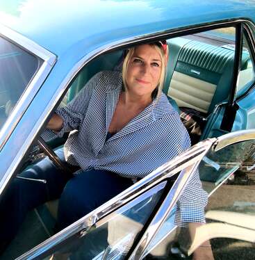 Uma mulher está sentada no banco do motorista de um carro azul antigo, sorrindo. Ela usa uma camisa xadrez e uma faixa vermelha na cabeça, com a luz do sol entrando.