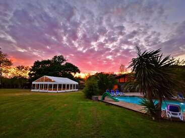 A vibrant sunset sky over a backyard with a swimming pool, lounge chairs, palm trees, green lawn, and a white event tent. Peaceful, relaxing atmosphere.