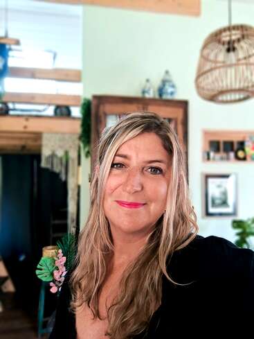 Uma mulher com longos cabelos loiros e um sorriso caloroso posa em um ambiente interno. Ela usa uma roupa preta com decorações florais, e o fundo é acolhedor e convidativo.