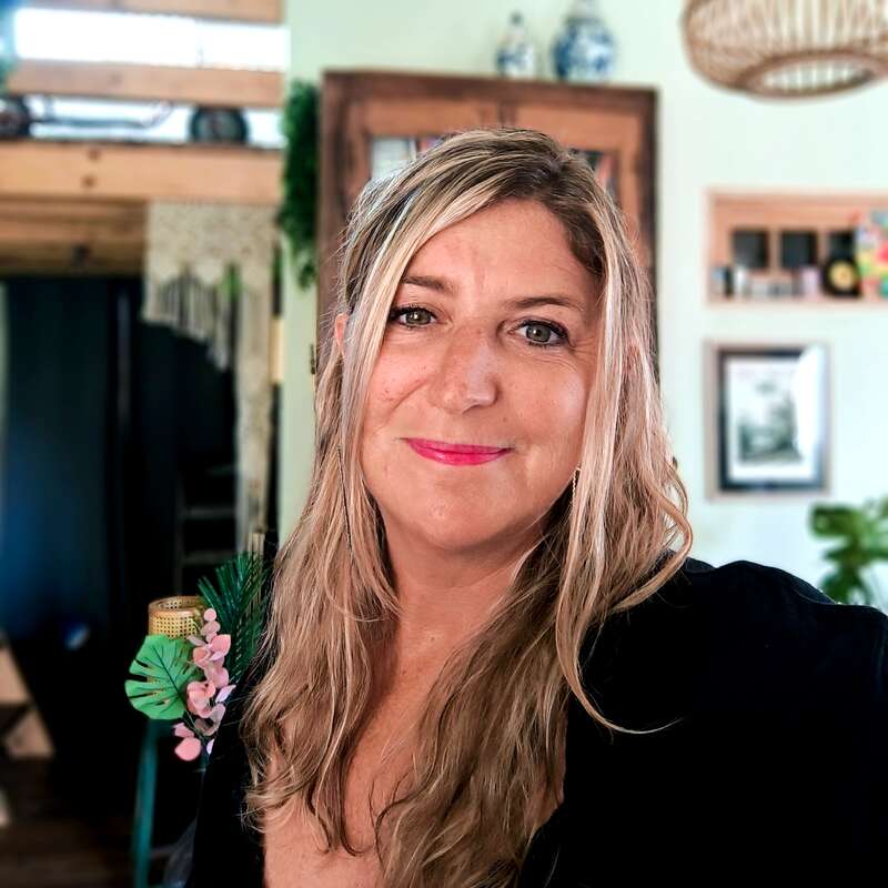 Uma mulher com longos cabelos loiros e um sorriso caloroso posa em um ambiente interno. Ela usa uma roupa preta com decorações florais, e o fundo é acolhedor e convidativo.